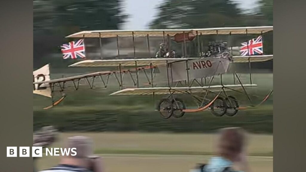An aircraft taking off as part of the Shuttleworth air show. There are flags on the plane and the word AVRO is on the side of it in red lettering.