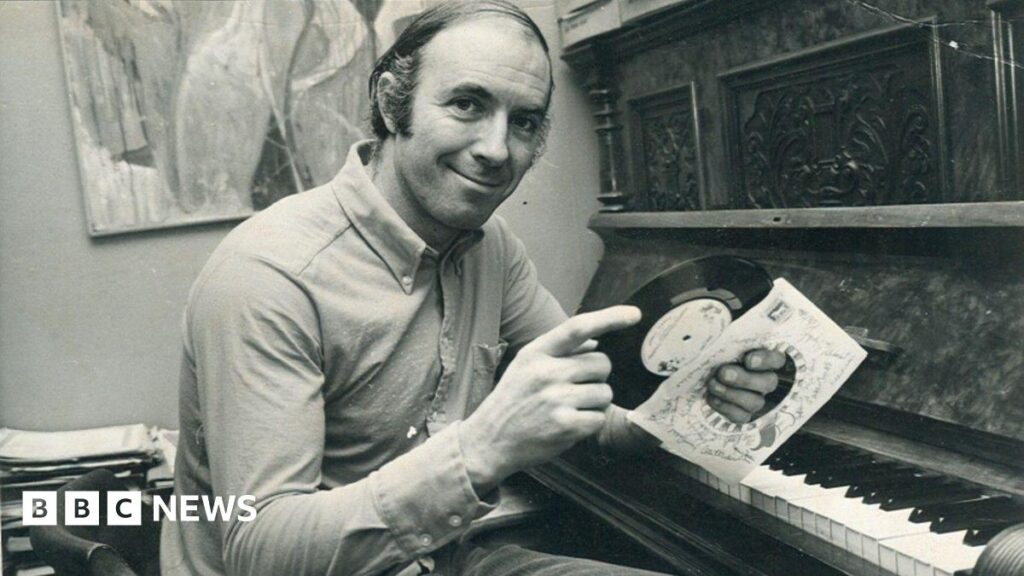 ‘We’re so proud of my dad and the Ipswich Town anthem he wrote’ A black and white image of a man sitting in front of a piano. He holds a small record in his hands and its sleeve. He has dark hair that has been slicked over one side of his head, and is wearing a shirt. He is smiling at the camera.