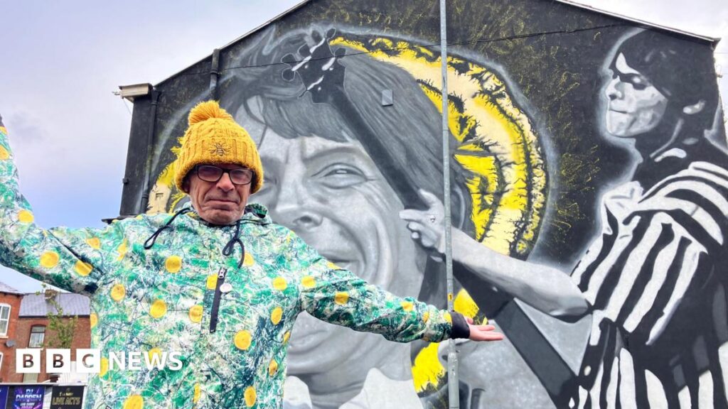 A man in his 60s wearing a yellow knitted bobble hat, glasses and a colourful anorak with lemons on the front stands in front of a large mural on the side of a building.