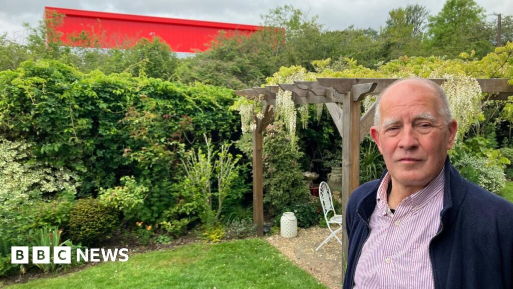 A man stands in a well-kept garden beside a wooden pergola with hanging white flowers, with dense greenery and a bright red building visible in the background under a cloudy sky.