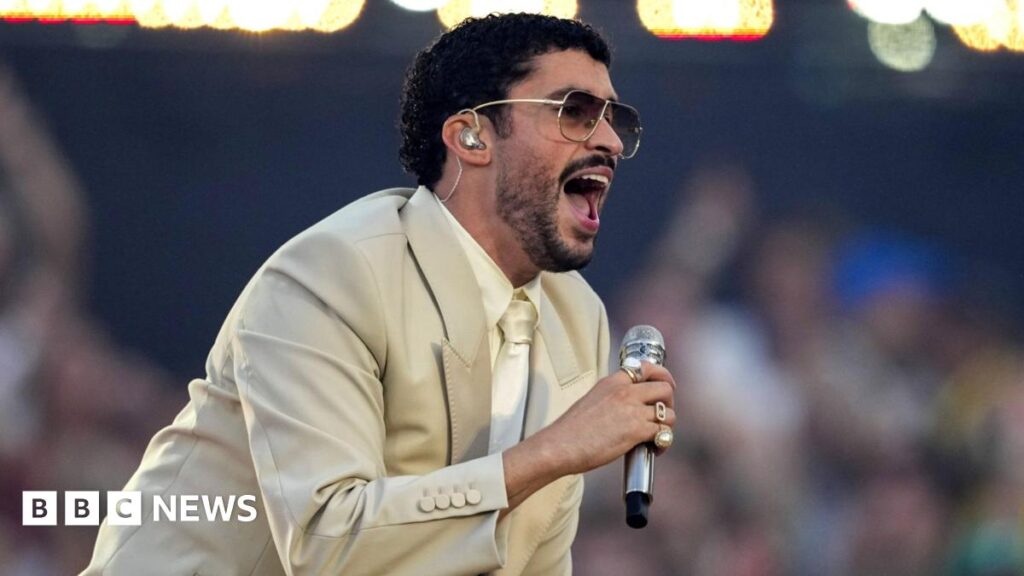 Bad Bunny to appear in Toy Story 5 as voice of Pizza With Sunglasses Bad Bunny in dark glasses, a beige suit, and cream shirt and tie singing into a microphone on stage during a concert in Barcelona on 22 May 2026
