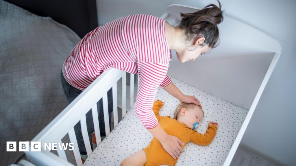 A woman leaning over a white cot, adjusting a baby lying on their back on a light-patterned mattress. The adult is wearing a red-and-white striped long-sleeved top, and the baby is dressed in an orange outfit. The scene is indoors, with a bed visible beside the cot.