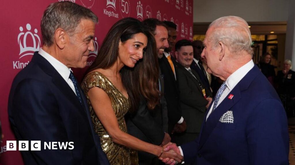Amal Clooney, dressed in a golden dress, shakes hands with a navy blue-suit wearing King Charles as her Hollywood star husband George, also in a dark blue suit, beams proudly at her right hand side during a line up of famous names meeting the monarch