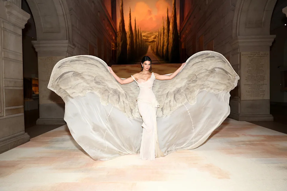 A person poses in an elegant gown with large wing-like extensions on the back, striking a dramatic and artistic pose inside a grand venue