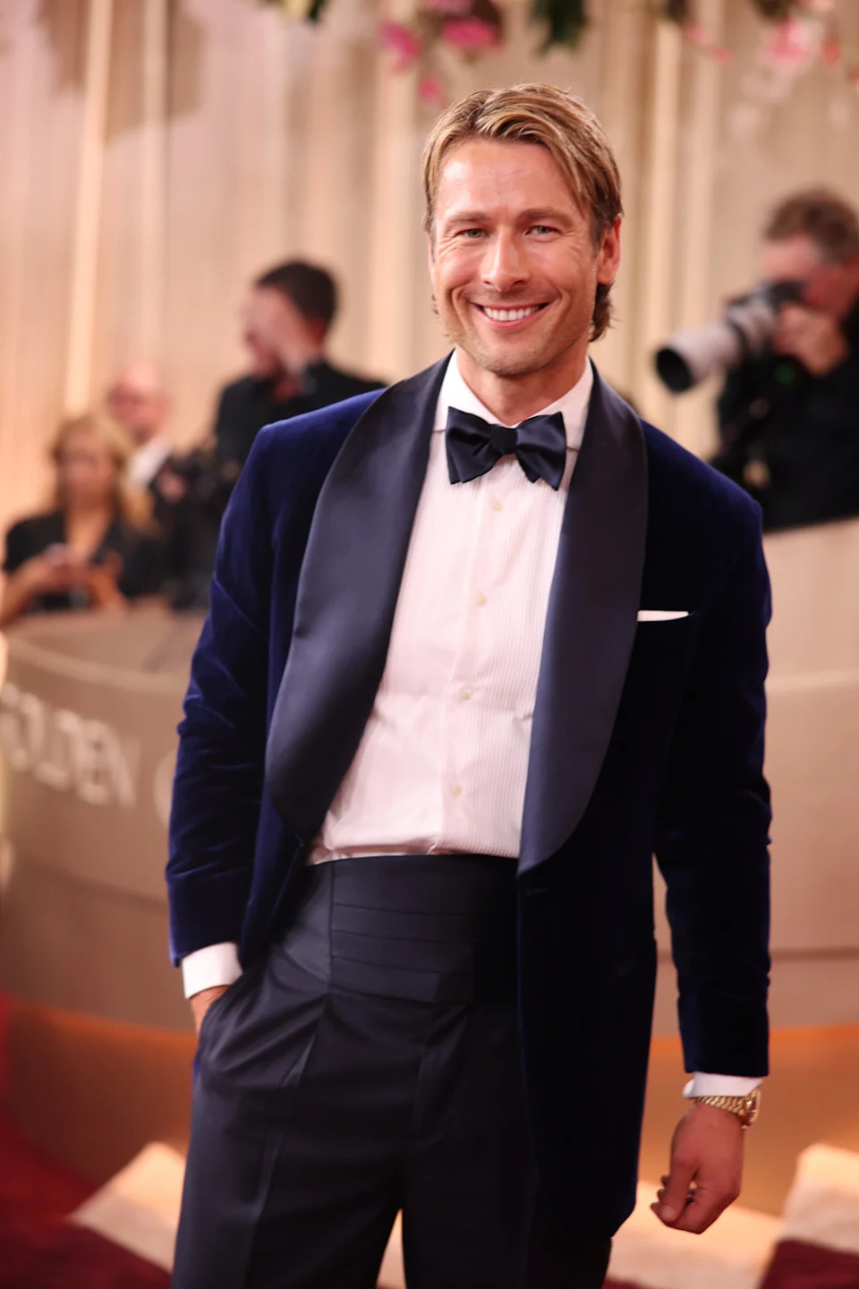 A person in a tuxedo with a velvet jacket poses and smiles on a red carpet event as photographers capture the moment in the background