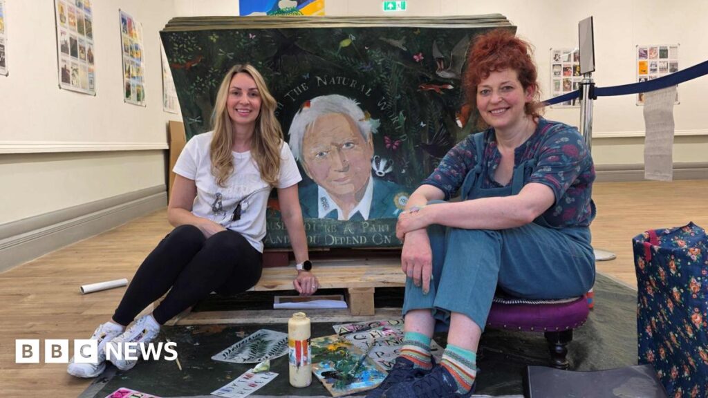 Suzy Hill has long blonde hair, white t-shirt and white trainers and black leggins. Lisa House has long red hair tied up, a patterned top, patterned socks, blue trousers and blue trainers. They are sat with the bench with art materials on the floor in a gallery setting.