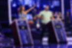 Jane waves beside Jamal on a purple-lit game show stage, with band behind them and podiums showing JANE $0 and JAMAL $4,000.