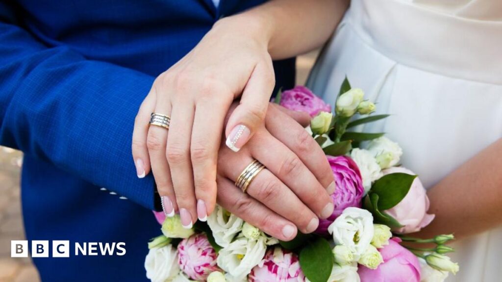 A stock image showing the hands of a bride and groom, resting on a bouquet of flowers and wearing wedding rings,