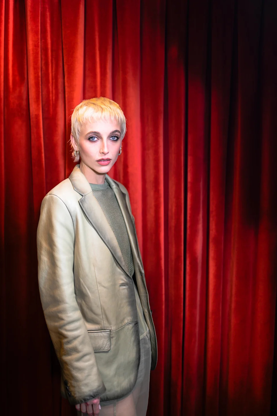 Person with short blond hair in a beige suit and mesh top stands against velvet drapes
