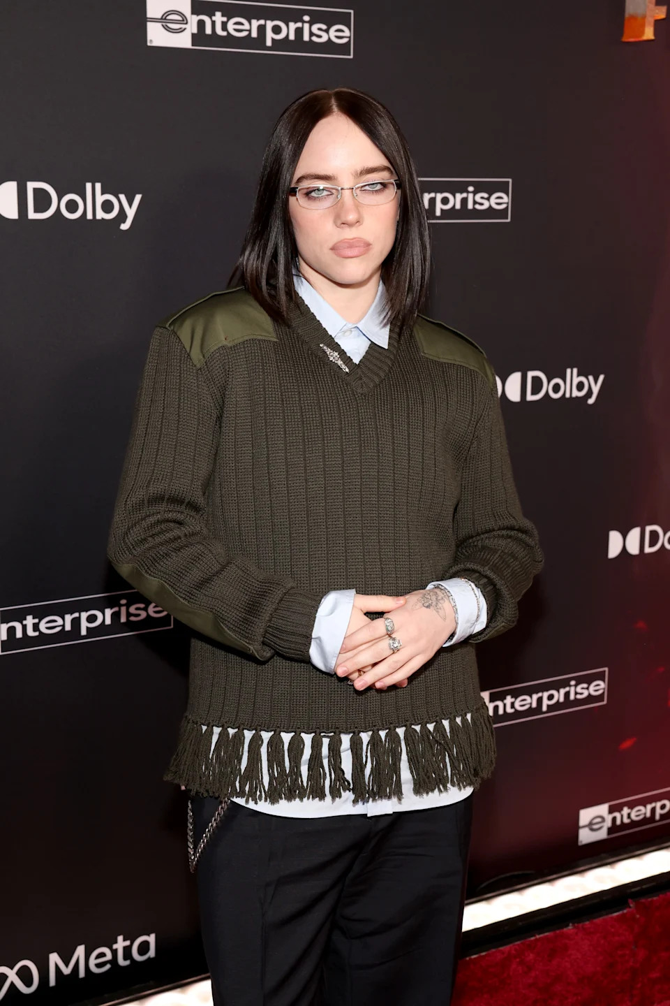 Person on red carpet wearing a fringed sweater over a shirt with chain details, and glasses, posing with hands clasped