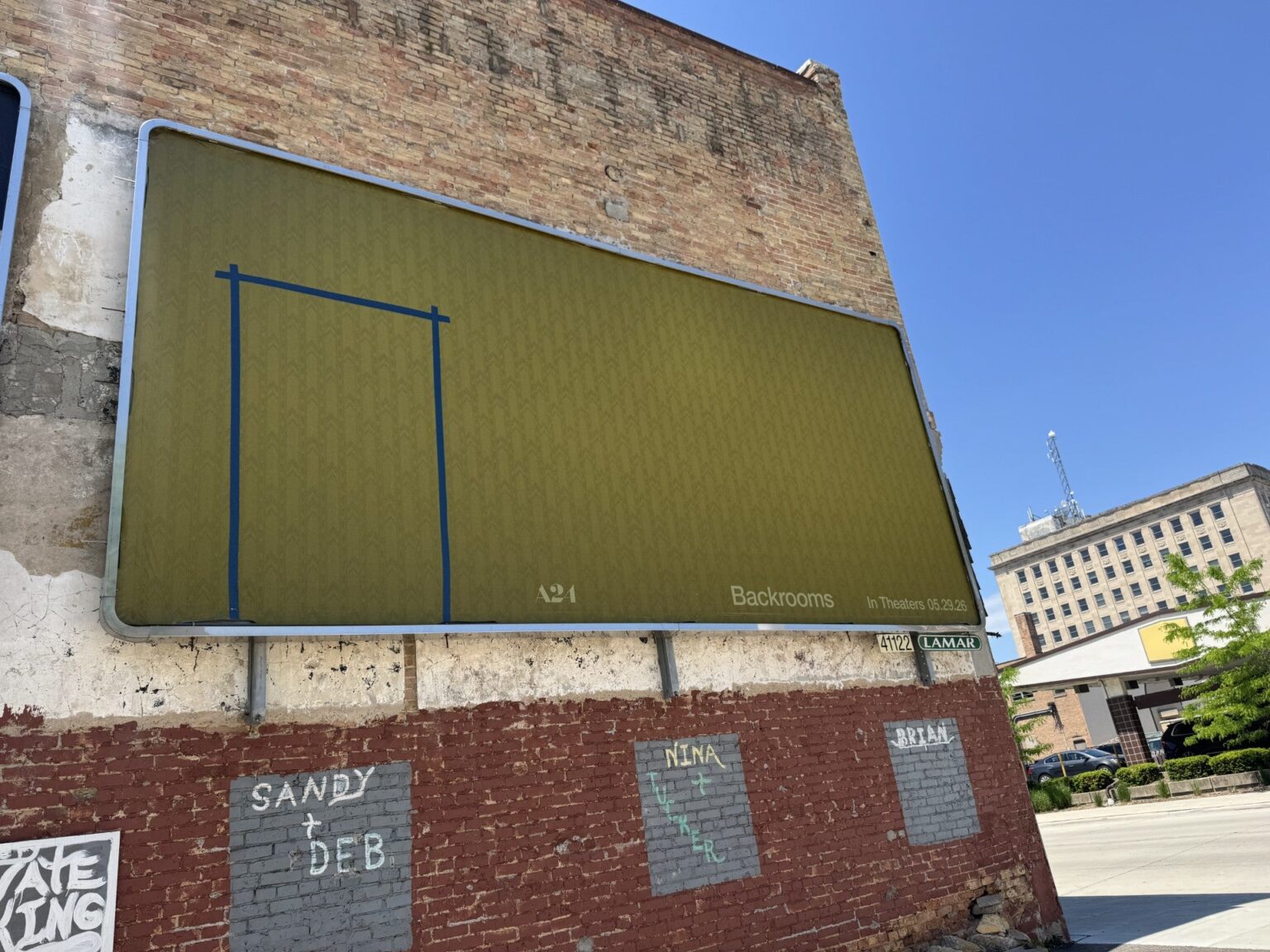 A yellow-green billboard with a faint Backrooms pattern and blue tape outline is mounted on a brick wall in an urban setting under clear skies.