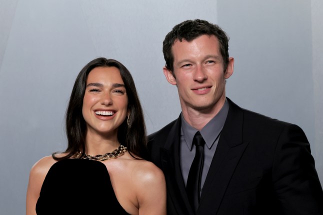 Dua Lipa and Callum Turner smiling together on a red carpet, in a black dress and black suit respectively.