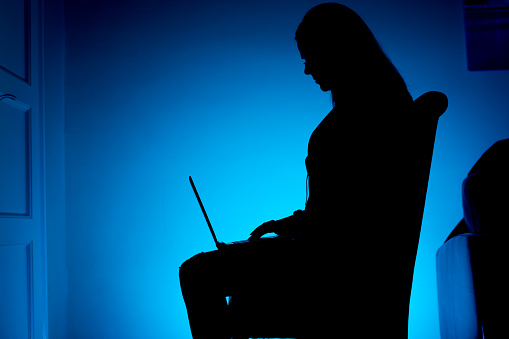 Silhouetted woman, sitting on the chair, working on laptop