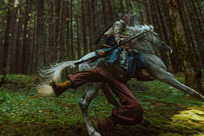 A man on horseback through the forest drags another man by the collar as he gallops.