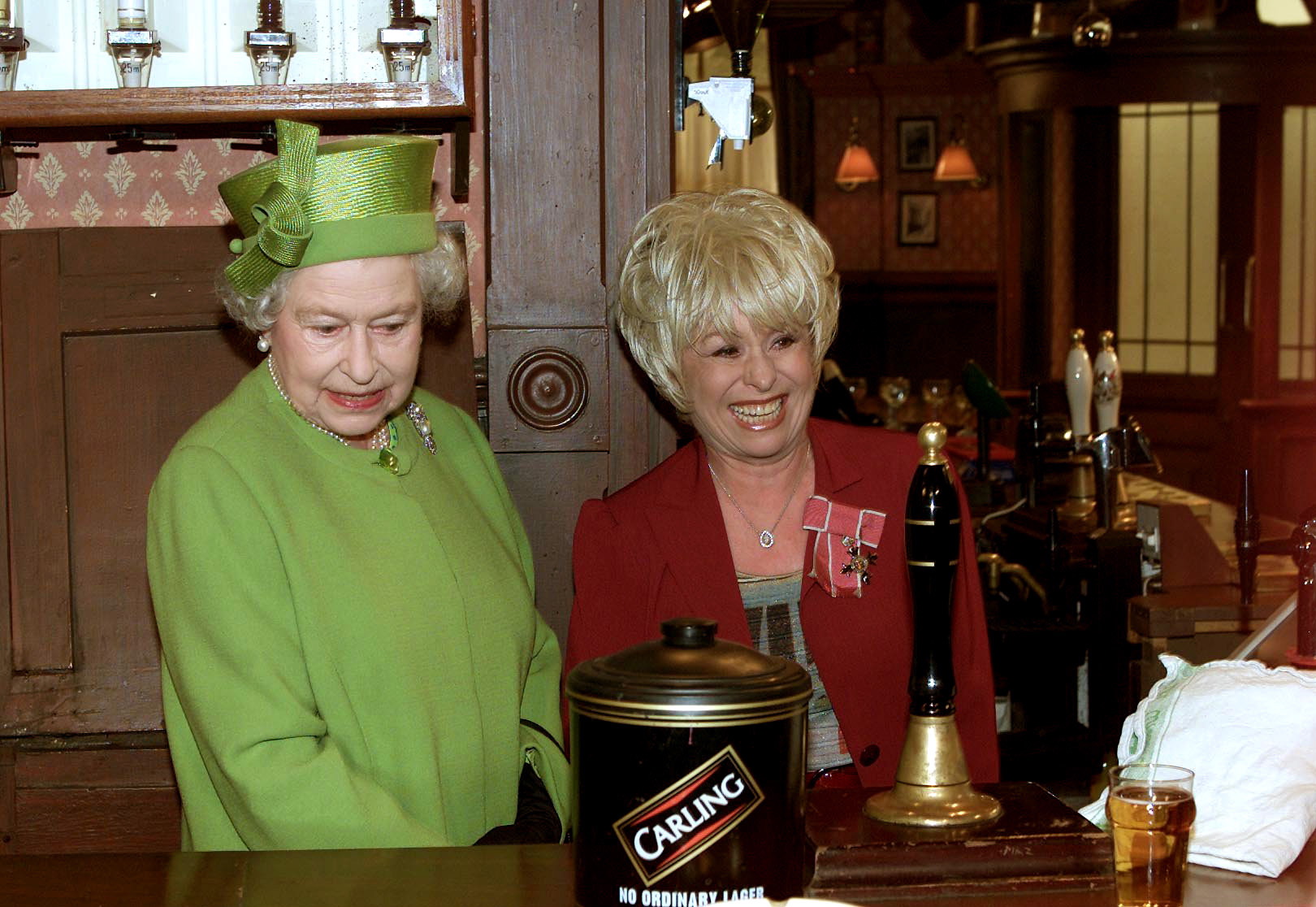 Queen Elizabeth in a green dress and hat behind the bar on the set of the soap opera Eastenders