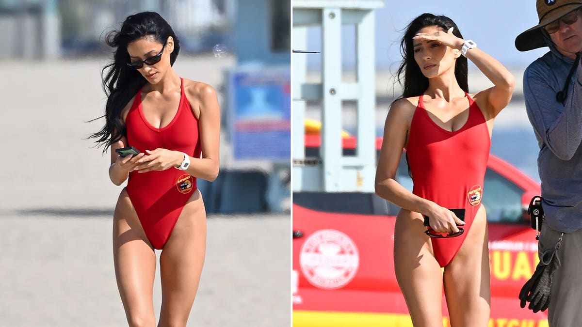 Shay Mitchell in red Baywatch swimsuit walking on Venice Beach set and shielding her eyes while filming