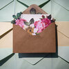 Photograph of a brown kraft paper envelope opened with paper flowers and lined paper bursting out. The envelope is elevated slightly above a background of green and yellow envelopes and the photograph is taken from above. .