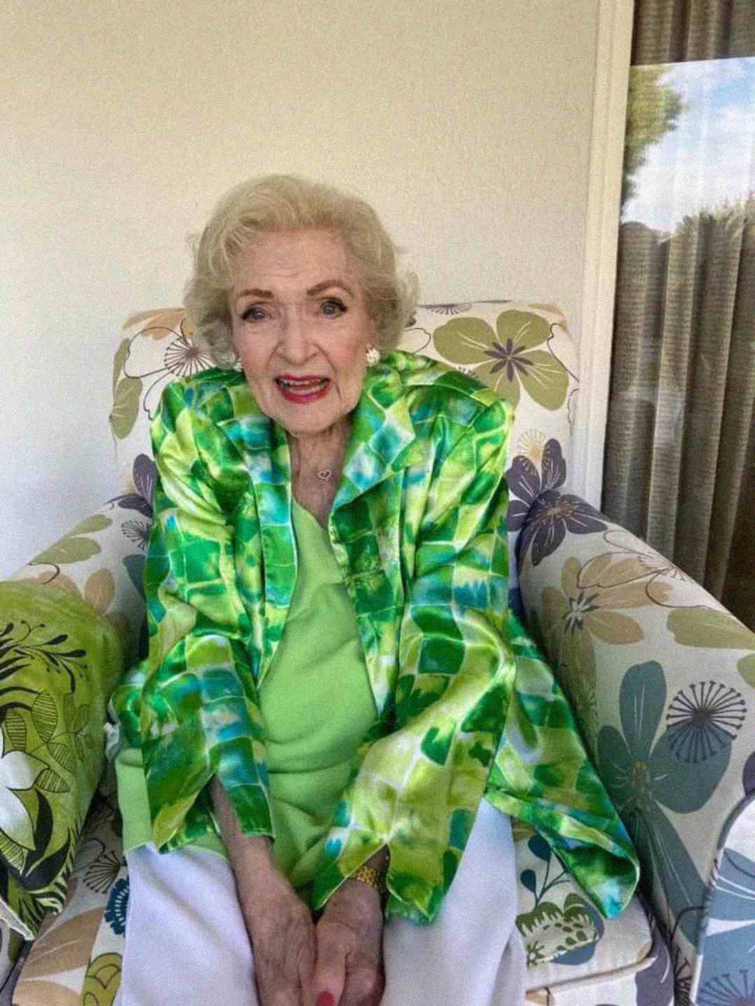 An elderly woman with white hair sits on a floral-patterned armchair, wearing a green and blue patterned jacket over a light green top and white pants. She is smiling gently at the camera.