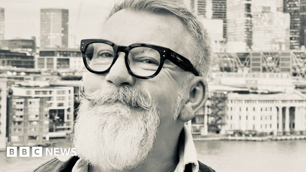 A black and white image of Simon Gee. He is smiling and wears dark-rimmed glasses. He has a distinctive moustache and beard.