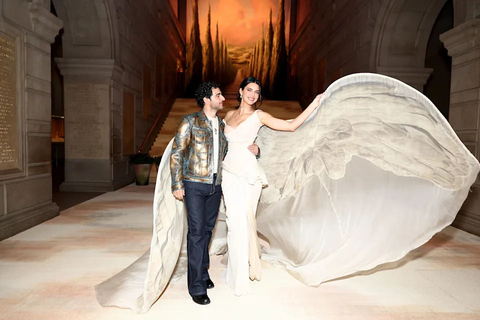 Two people pose indoors; one wears a metallic jacket, and the other wears an elegant, flowing gown with large fabric wings