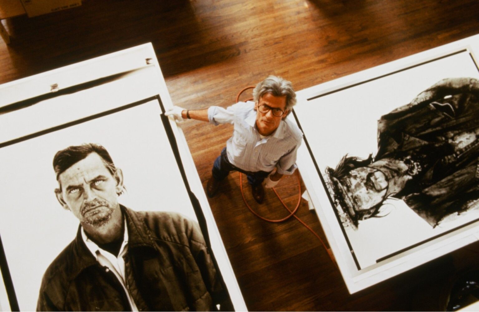Shot from above, Avedon stands on a wooden studio floor between two enormous exhibition prints from his In the American West series, dwarfed by the faces around him.