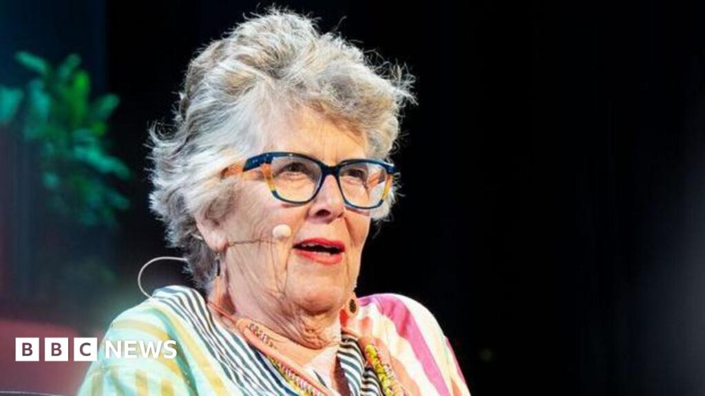 A man and a woman sat on a stage with crowds in front of them. There is a small table with water in front of them. The man has short brown hair and is wearing a grey t shirt and glasses. The woman is wearing a multicoloured stripy dress and glasses and has short grey hair. Behind them is a multicoloured background, plants and the Hay festival logo.