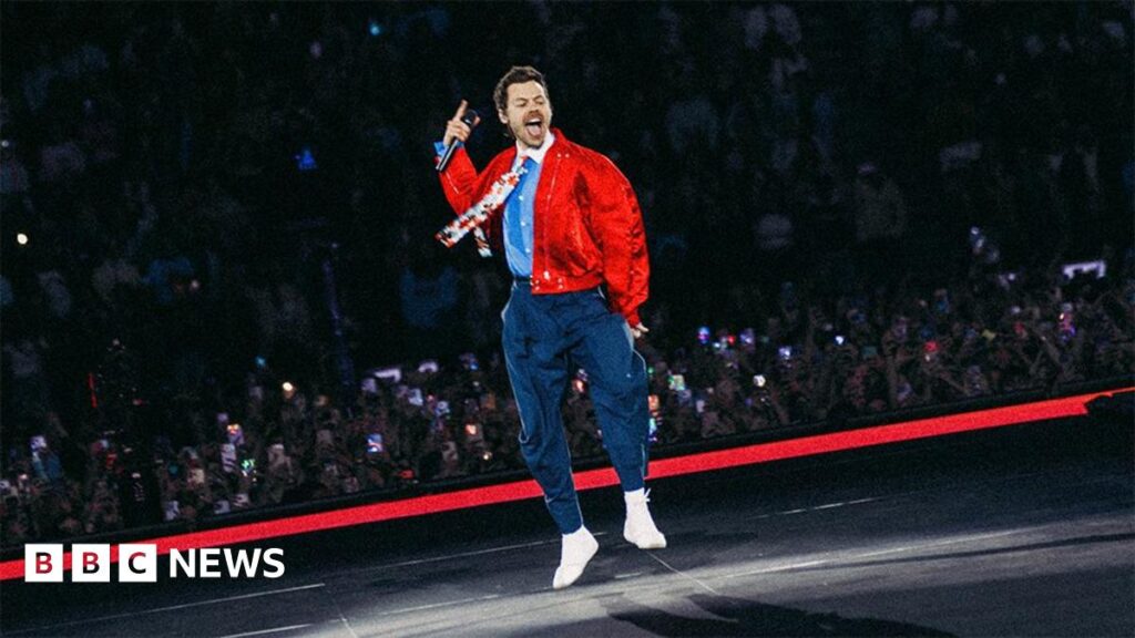 Harry Styles tour to review staging after fan complaints Harry Styles is wearing navy trousers with a belt and a light blue shirt with a white collar and flower patterned tie.He has a red bomber jacket over the top. He is jumping on the stage, you can see fans watching from the crowd behind him.