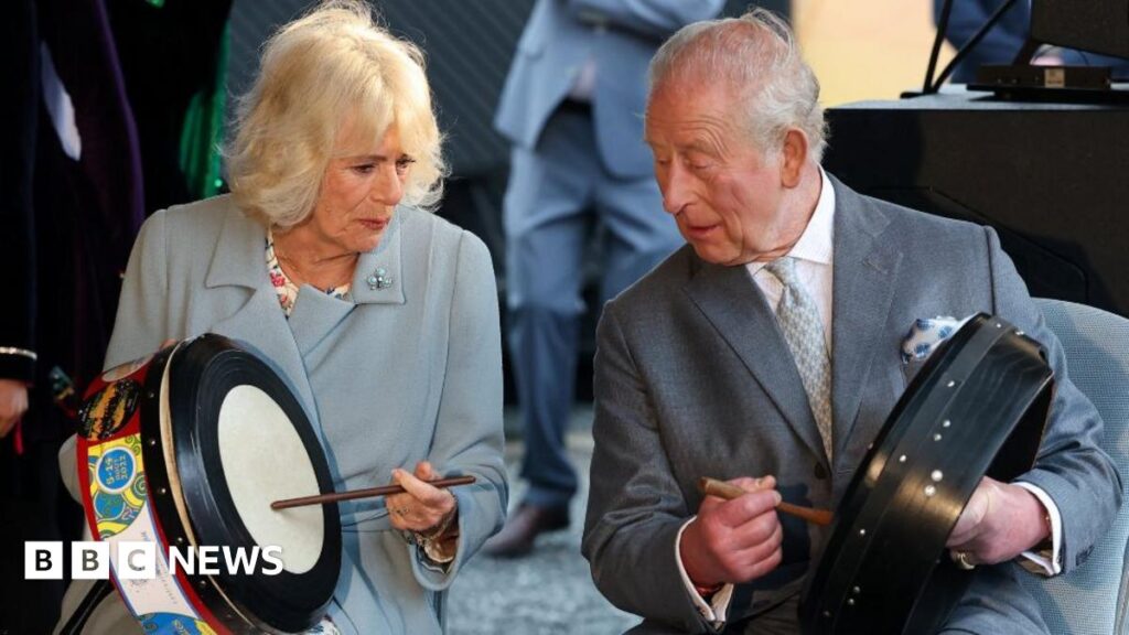 King Charles smiles and waves. He is wearing a grey suit and pale grey tie. Queen Camilla stands beside him wearing a long blue coat and she carries an umbrella. There are two men in suits either side of them in the photo. Photographers in the background as well as signage for the Fleadh.