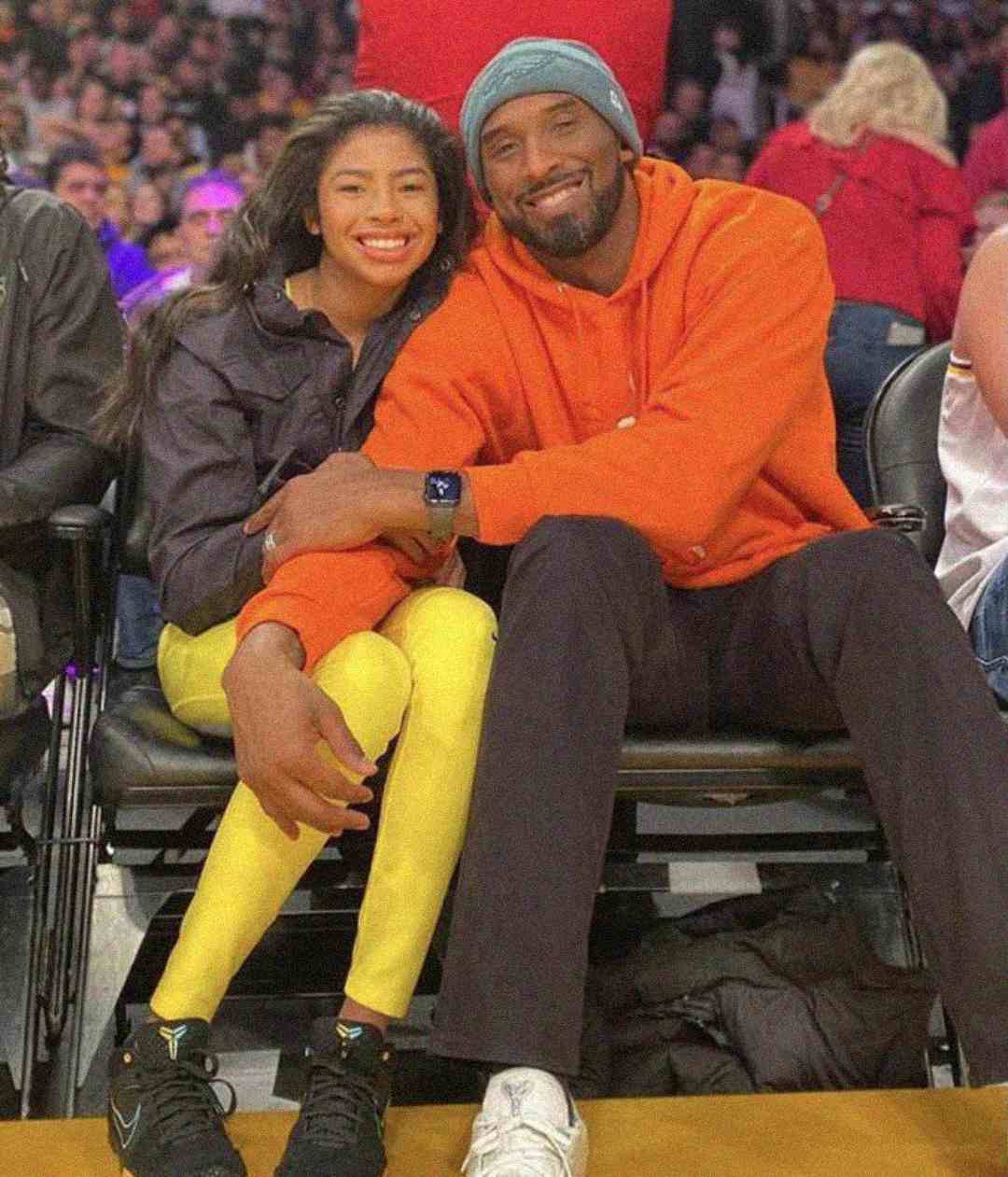 A smiling man in an orange hoodie and a young girl in a black jacket with yellow pants sit closely together courtside at a basketball game, surrounded by a cheering crowd.