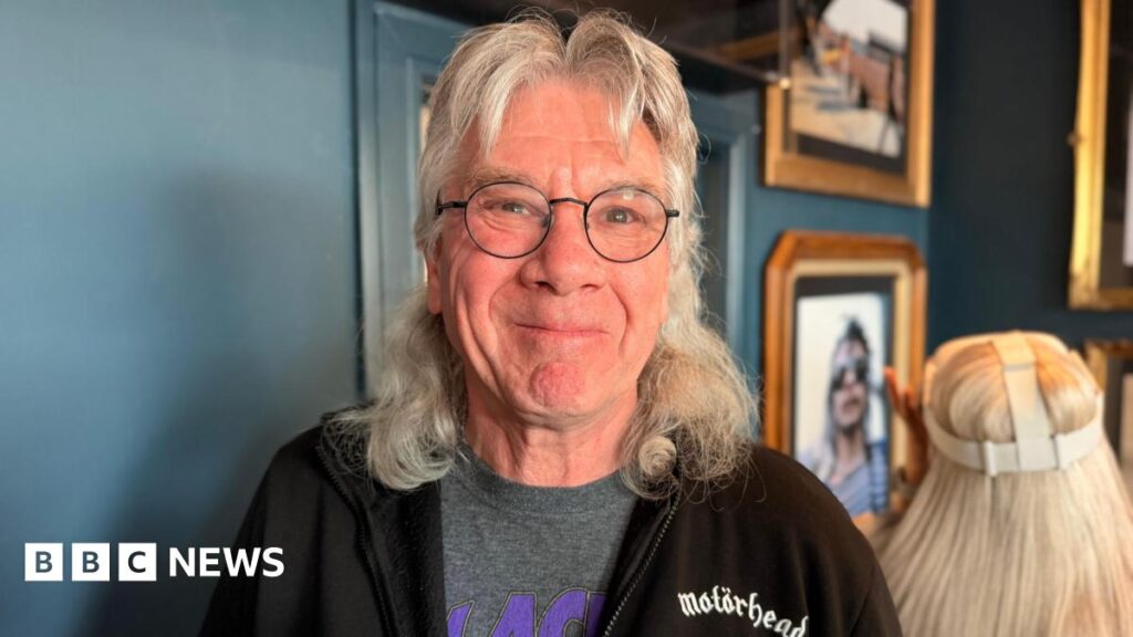 A man with long grey hair cut into a mullet style. He has black rimmed round glasses and is wearing a black Motorhead hoodie with a grey and purple Black Sabbath t-shirt underneath.