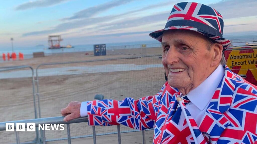 An elderly man pictured in front of a beach wearing a suit covered in Union flags and wearing a hat with the same pattern on it.