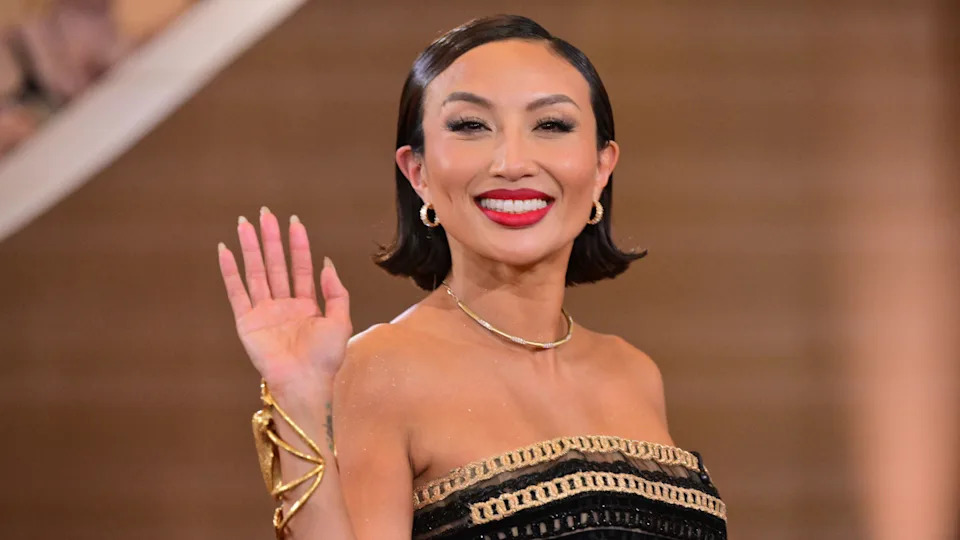 Jeannie Mai with short hair, wearing a strapless black and gold dress, waves and smiles at the camera.