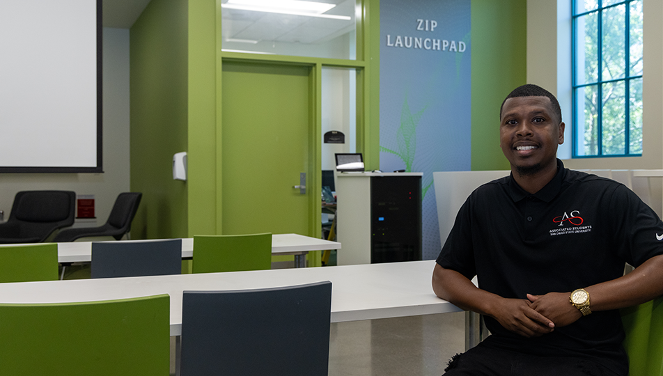A young man in a black polo shirt sits with his right elbow on a desktop in a room marked ZIP LAUNCHPAD on the wall.
