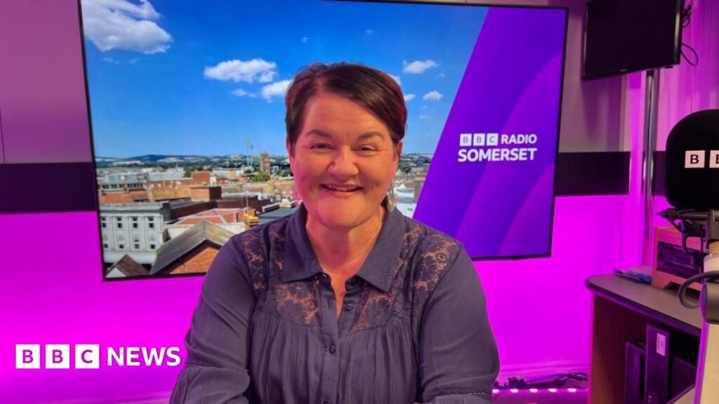 Emma Britton in the BBC Radio Somerset studio. She has short dark hair, is wearing a blue top in a purple radio studio with a screen and a microphone.