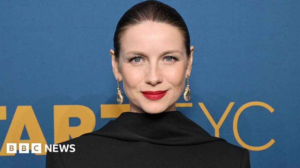 US site apologises to Outlander star over Shotts ‘armpit’ misquote Caitriona Balfe, with her hair slicked back and wearing visible makeup including striking red lipstick as well as long jewelled earrings. She is standing in front of a blue sign with yellow writing on it.