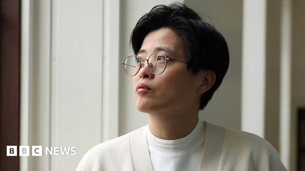 Yang Shuang-zi has short dark hair, wears thin, black rimmed glasses and wears a cream cardigan over a white t-shirt. Standing next to her, Lin King has slightly longer, shoulder length dark hair, and wears a blue and white striped shirt.