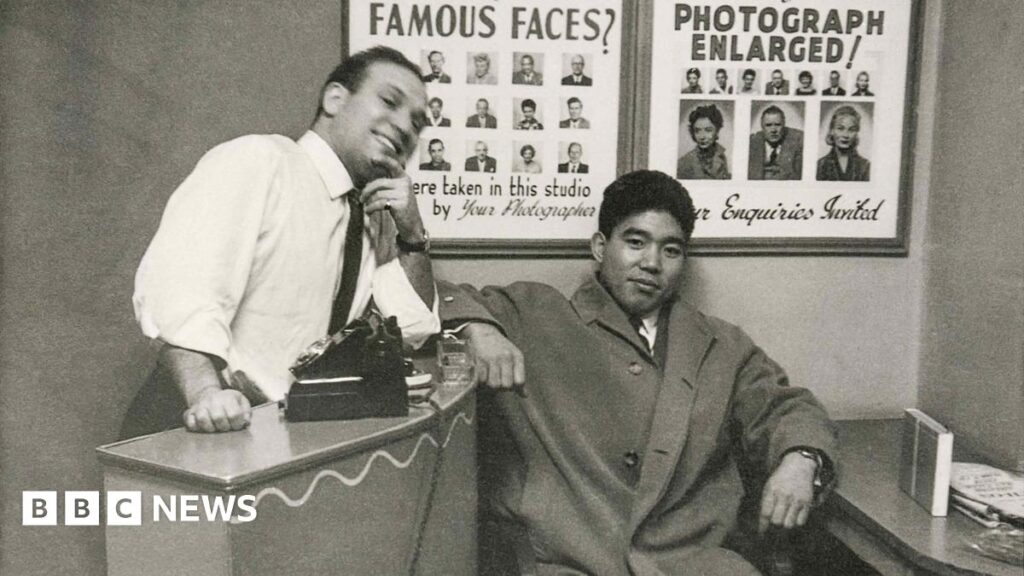 Dave Sharkey, with customer, 1962. Black‑and‑white photograph of Dave Sharkey, with customer. Dave leans against a counter with a camera placed on top, while the other sits beside the desk. Posters of sample portrait photographs hang on the wall behind them.