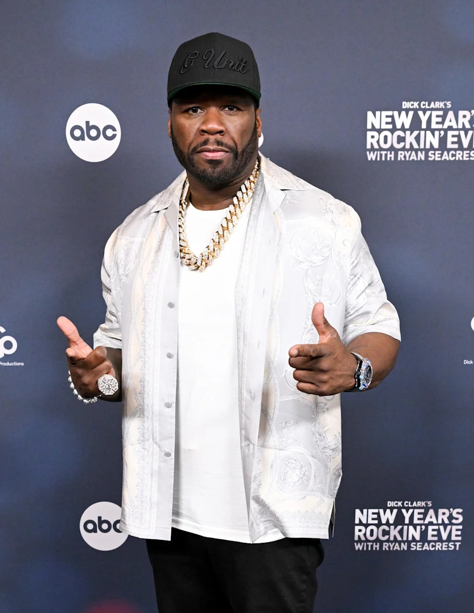 A person in a patterned shirt, white T-shirt, cap, and gold chain poses at a "New Year's Rockin' Eve" event backdrop