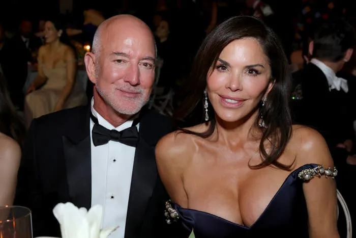 A couple in formal evening attire at a gala event; the man in a tuxedo, the woman in an elegant off-shoulder gown, sitting together