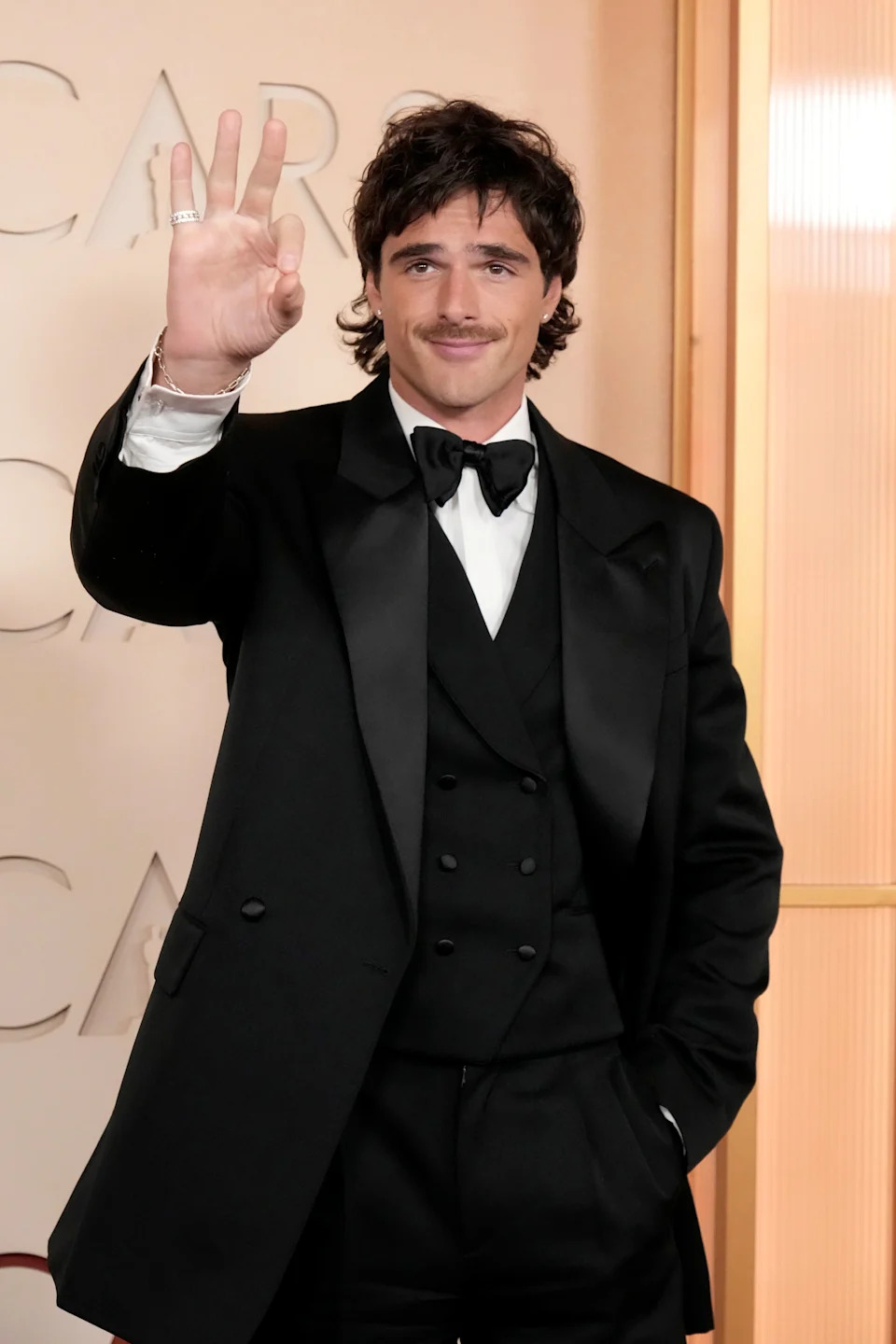 Person in a formal black tuxedo with a bow tie, posing with a hand gesture on a red carpet event backdrop