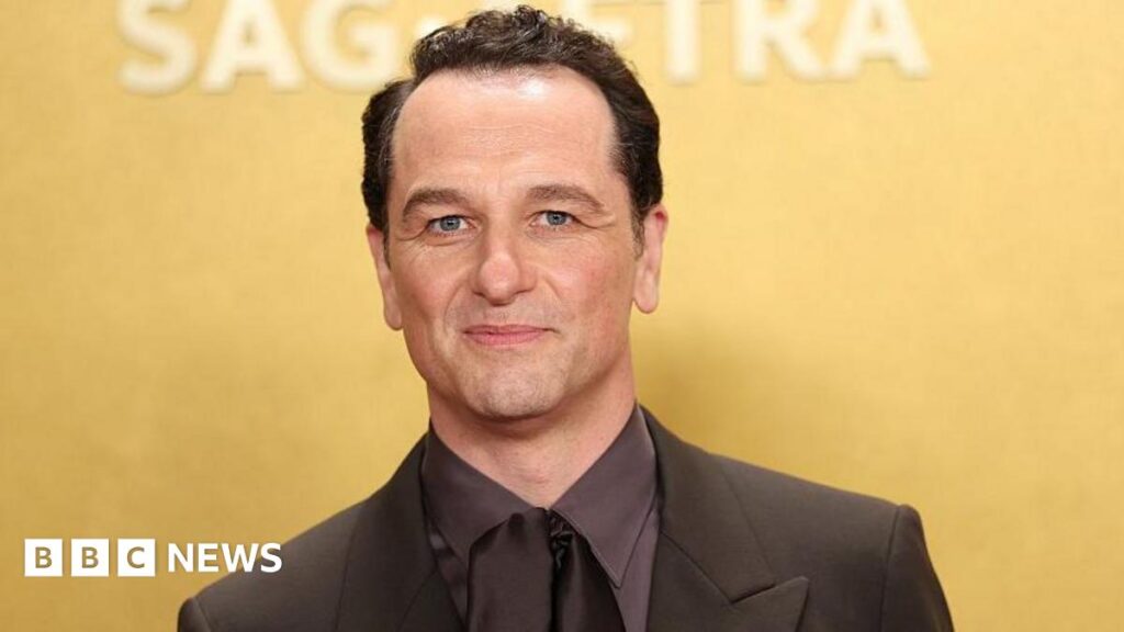Matthew Rhys in Widow's Bay. He is sat down, holding a folder, looking straight ahead