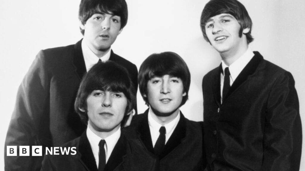 A black and white photo showing the four members of The Beatles when they were in their 20s, all wearing matching black suits, white shirts and thin ties, smiling into the camera.
