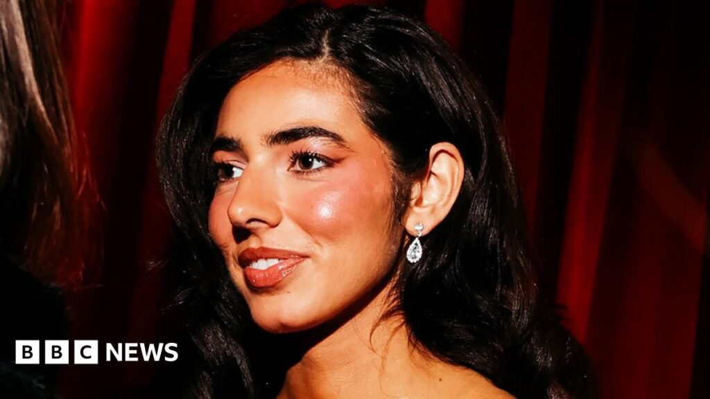 Izzy Escobar wears a black strappy formal dress with black gloves on the red carpet of the Devil Wears Prada 2 premiere. She has long wavy black hair and wears a silver necklace. She stands against a backdrop showing the film title