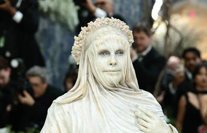Person in creative, draped outfit and headpiece, posing at a formal event amid photographers