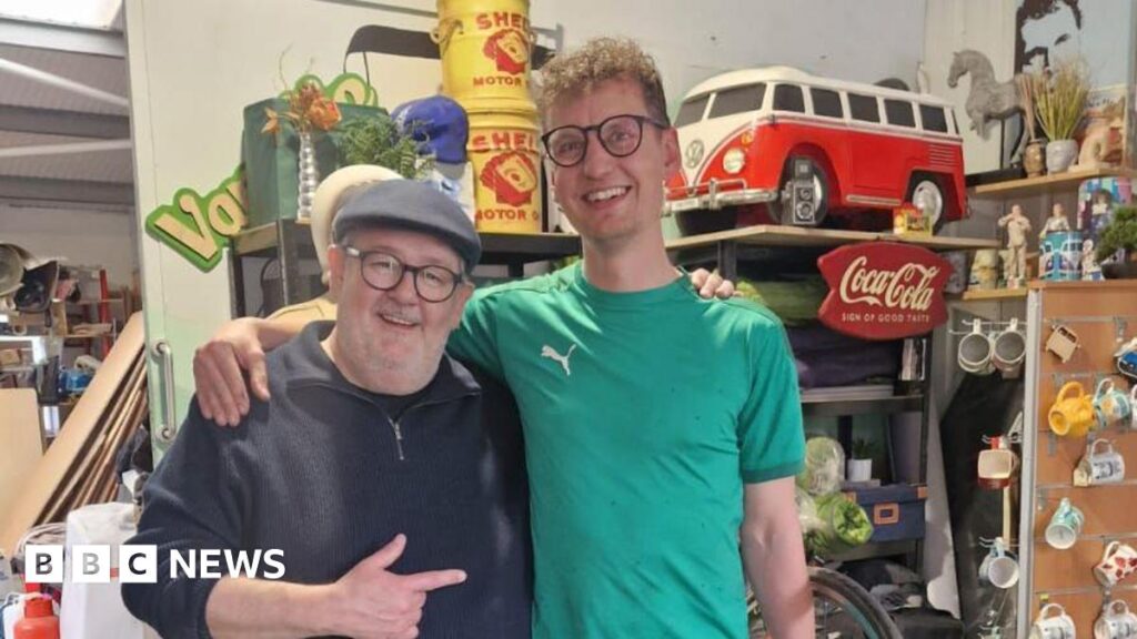Two men in glasses, each with an arm around the shoulder of the other. They are both smiling, the older man is wearing a flat cap