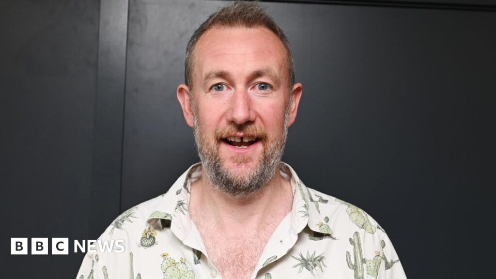 Taskmaster creator Alex Horne launches hospital club for child patients The image shows a man making a slightly shocked expression as he looks at the camera. He is wearing a beige shirt with cacti on it.