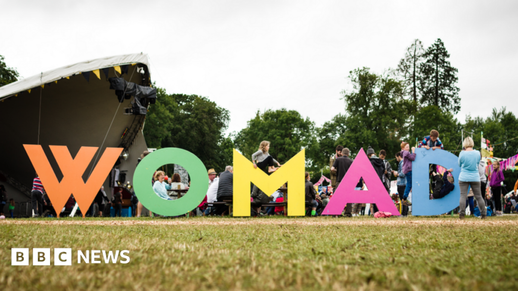 A field with a large music stage in the background and lots of people in front of it. In the foreground of the picture there are huge multi-coloured letters spelling out 'WOMAD' in capital letters