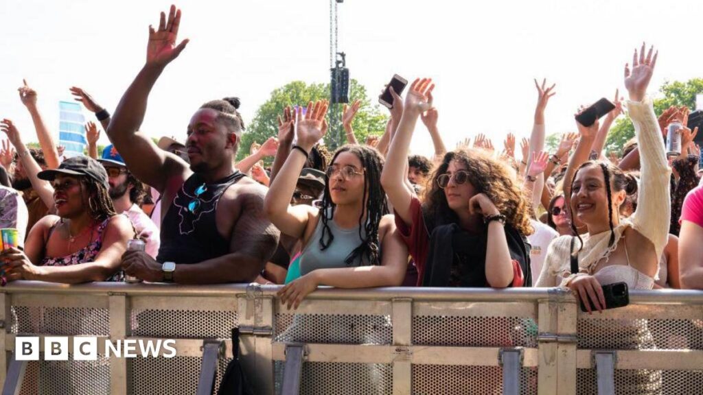 Crowd of festivalgoers standing behind a metal safety barrier at an outdoor music event, many with their hands raised in the air. A group at the front includes several young adults leaning on the barrier, some holding drinks and mobile phones, with trees and stage equipment visible in the background under bright daylight.