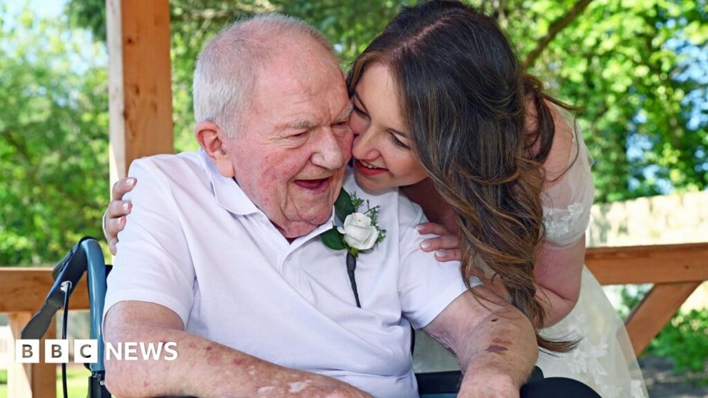 Cardiff bride has two weddings in two days for grandad with dementia A woman in a wedding dress on the right smiles as she looks at a man. The elderly man is wearing a white roses pinned to to his top and is sitting in a wheelchair with the handle visible on the left.
