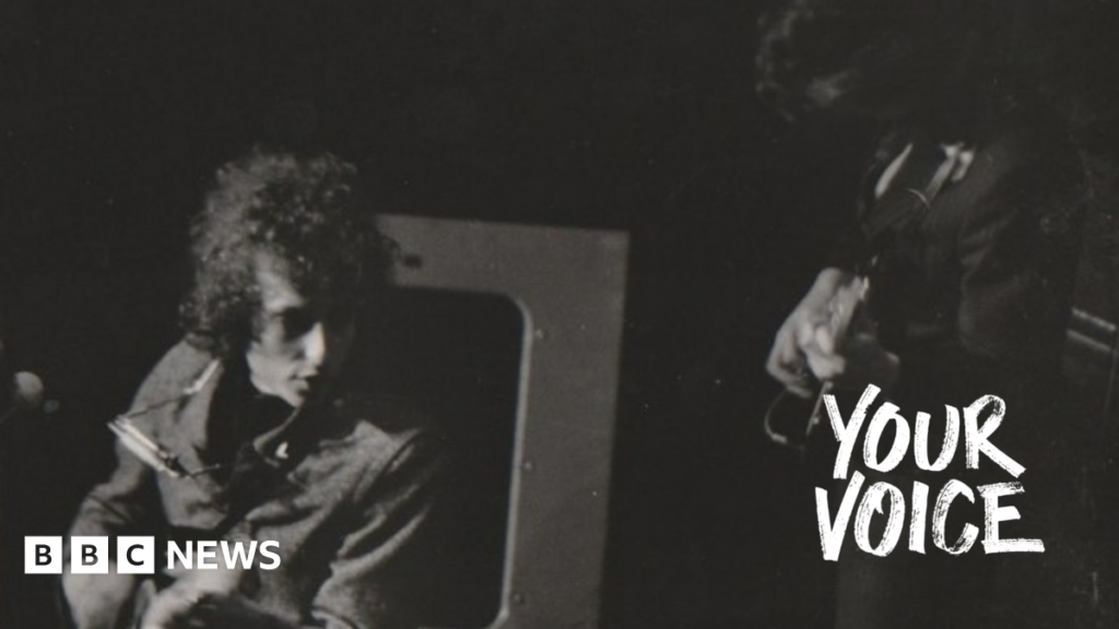 Bob Dylan with his distinctive curly hair, has his harmonica round his neck and is playing his electric guitar at Manchester Free Trade Hall